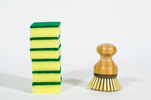 A stack of yellow and green sponges is sitting on a white background. Next to it is a wooden dish brush.
