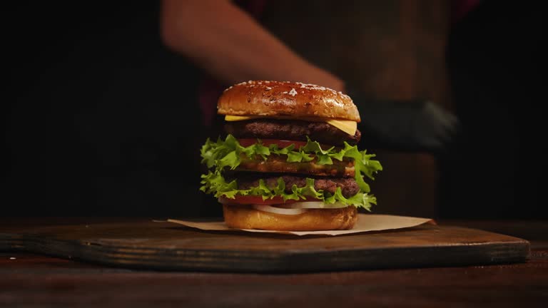 Chef making burger on serving board. Cheeseburger with beef patty, cheese, tomato, onion, fried bun, lettuce, sauce