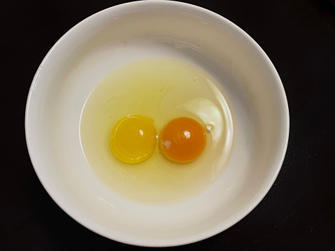 Two cracked eggs with no eggshell in a white bowl.   Black background.   One orange yolk egg.  One yellow yolk egg.