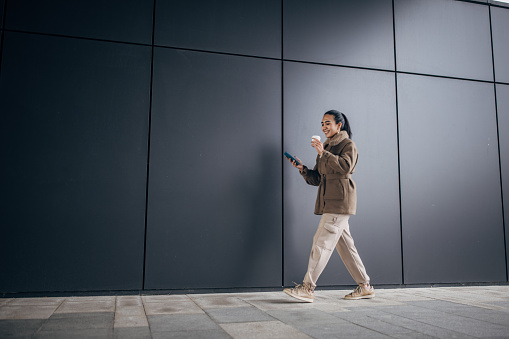 One woman, Asian woman walking on sidewalk outdoors, she is holding a coffee cup and using smart phone.