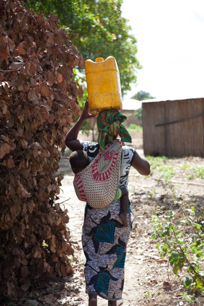 las mujeres recogiendo agua en cubos y llevando balanceada sobre la cabeza con el niño en una capulana o portabebés o caminando al lado. - baby child poverty water fotografías e imágenes de stock