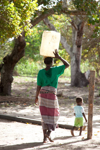 las mujeres recogiendo agua en cubos y llevando balanceada sobre la cabeza con el niño en una capulana o portabebés o caminando al lado. - baby child poverty water fotografías e imágenes de stock