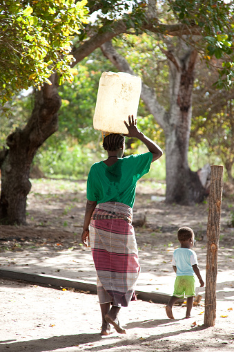 Xai Xai Mozambique: Women collecting water in buckets and carried balanced on head with child in a capulana or baby carrier or walking beside.