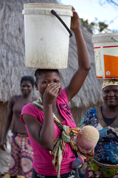 las mujeres recogen agua en cubos y llevan equilibrada en la cabeza con el niño albino en una capulana o portabebés o caminando al lado. - baby child poverty water fotografías e imágenes de stock