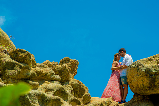 engaged couple next to a rocky area and a blue sky with their faces close together and an embrace.