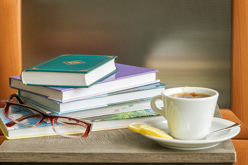On the table are a stack of books to read, women's glasses for vision, a cup of hot tea for a pleasant pastime at home.