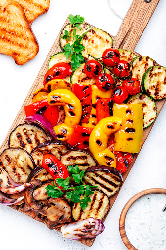 Grilled vegetables: red and yellow paprika, zucchini, eggplant, mushrooms, tomatoes and onions served on rustic wooden cutting board, white table background, top view