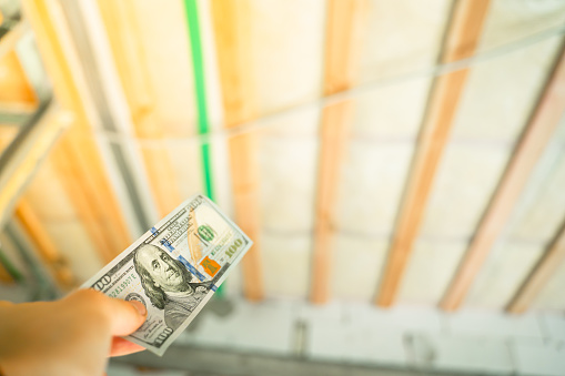 A hand holds a hundred dollar bill against the background of the insulated ceiling of a private house. payment for internal work during repairs in the house