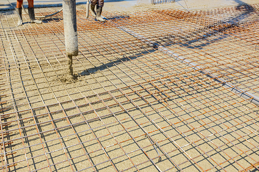 Pump hose is pouring fresh concrete over square reinforcement in the base of new building covering sanitary pipeline, set of embed plastic pipes for sewer.