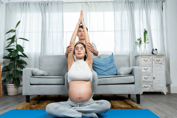 un homme et une femme asiatiques sont assis sur un tapis de yoga dans le salon d’un poste de police. un père qui aide une femme enceinte avec un gros ventre essayez le yoga pour vous détendre dans les jours précédant l’accouchement. - torso physical therapy patient relaxation exercise photos et images de collection