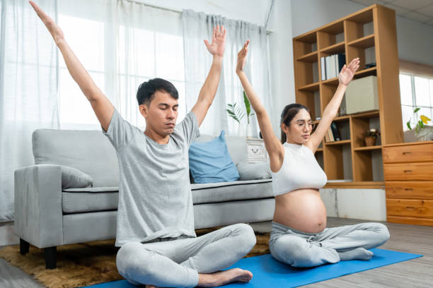 아시아 남성과 여성은 배꼽 셔츠를 입은 임신한 아내를 둔 남편과 아내입니다. 거실 한가운데서 여유롭게 휴식을 취하기 위해 머리 위로 손을 들고 요가 자세로 앉아 있습니다. - torso physical therapy patient relaxation exercise 뉴스 사진 이미지