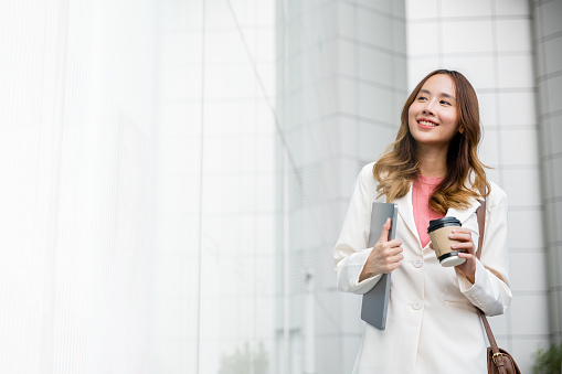 Business woman wearing white suit jacket and smile while go to work outdoor mirror building background. Confident Businesswoman with a cup of coffee and holding Laptop walking outside office building