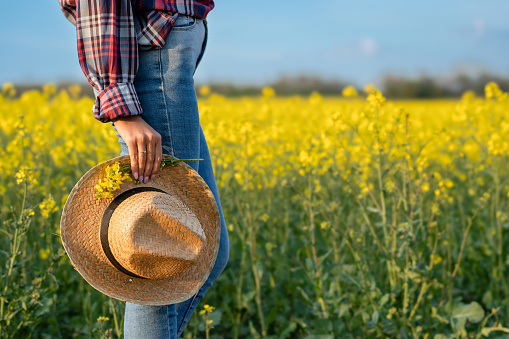 Side view of female farmer standing in blooming rapeseed field and holding straw hat. Farming and agriculture concept