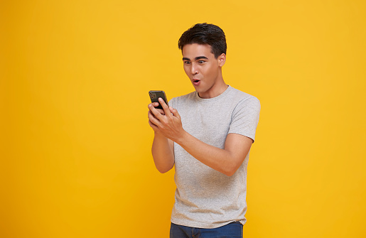 Surprised young asian man using mobile phone with positive expression isolated on yellow background. Happy wow glad man success.