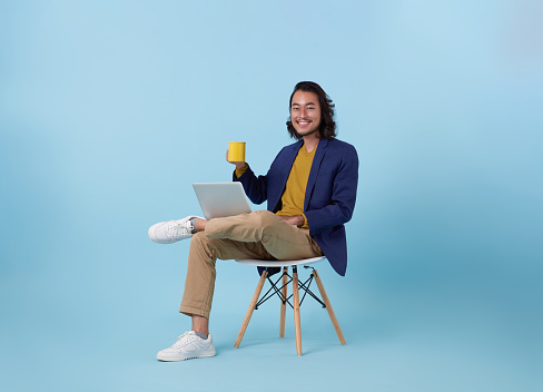Young asian business man happy smiling using computer laptop sitting on white chair and drinking coffee isolated on blue studio background.