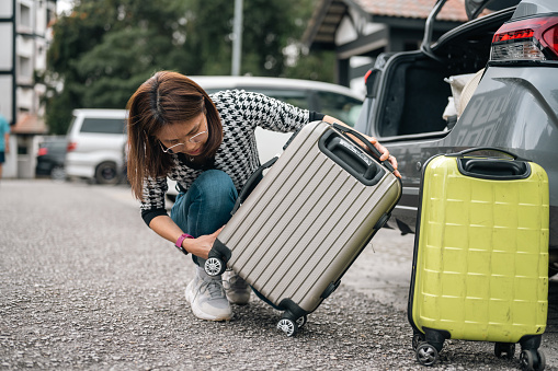 An Asian woman is checking her travel suitcase, which has a broken wheel, while traveling by car.