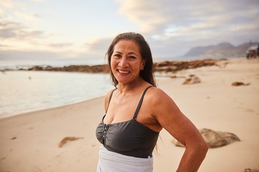 Portrait of a mature woman wearing a swimsuit and standing wrapped in a towel smiling on a beach after an ocean swim