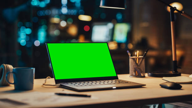 istock Laptop Standing on a Desk with a Green Screen Chromakey Mock Up Display. Table with Computer, Coffee Mug, Headphones and Notebook in Creative Office at Night. 1481145918