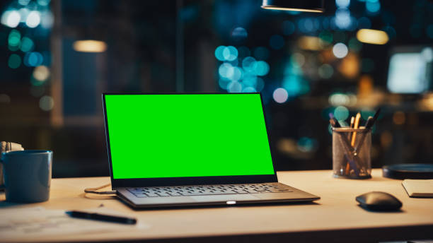 istock Laptop Standing on a Desk with a Green Screen Chromakey Mock Up Display. Table with Computer, Coffee Mug, Headphones and Notebook in Creative Office at Night. 1481145923