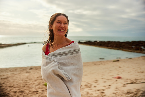 Laughing mature woman wearing a swimsuit standing wrapped in a towel on a beach after going swimming in the ocean