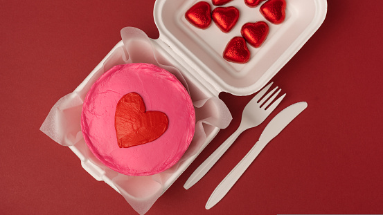Valentine day bento cake In eco box with knife and fork and heart shaped candies on red background, flat lay
