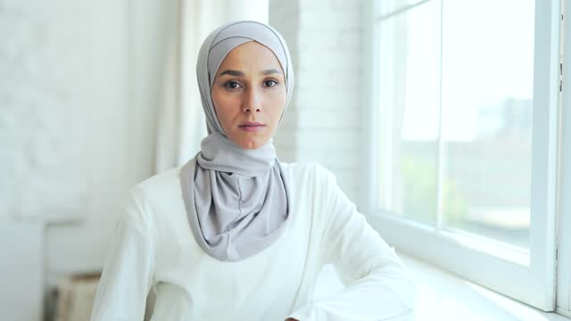 closeup portrait a serious confident young Muslim woman in a hijab standing indoors at home