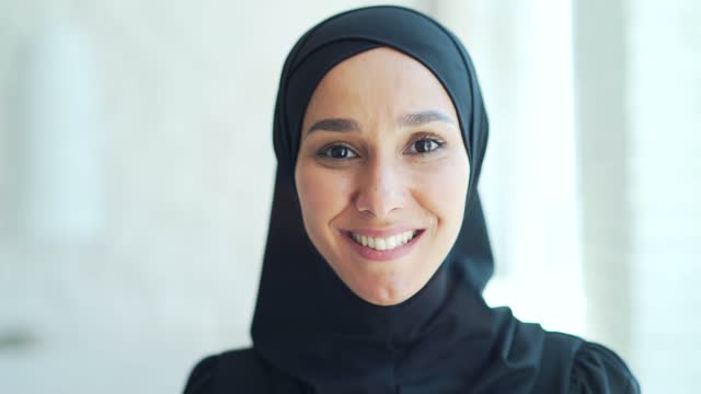 closeup portrait of a pretty young Muslim woman in a black hijab standing and smiling indoors at home Headshot of an Islamic happy female looking at the camera