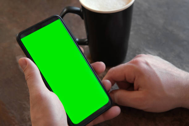 Man Use of Mobile Phone with Blank Green Screen in Cafe on Rustic Vintage Wooden Table. Male Holding Smart Phone Empty ChromaKey Screen Mock up in Coffee Shop. Morning. Showing Mock up Man Use of Mobile Phone with Blank Green Screen in Cafe on Rustic Vintage Wooden Table. Male Holding Smart Phone Empty ChromaKey Screen Mock up in Coffee Shop. Morning. Showing Mock up. Young man hand use smartphone with green screen. Gestures pack. Male hand touching, clicking, tapping swiping on black phone chromakey display. Close up Device. Modern Technology. Internet social media. Caucasian Man finger. Guy using phone mockup. Close up. Using for a smartphone, tablet pc or a touch screen devices. Gestures. cellphone with tracking markers. watching content. modern technology man looking at phone over shoulder stock pictures, royalty-free photos & images