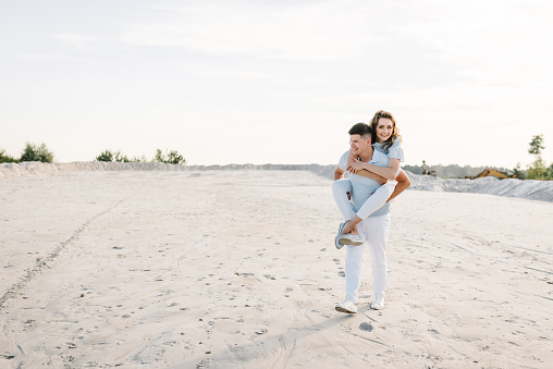 A handsome young man giving a piggyback ride to a woman in summer sunset. Girl sits on back and hugs boyfriend on the sand on the sandy beach. Lovely couple having fun. Time spent together.