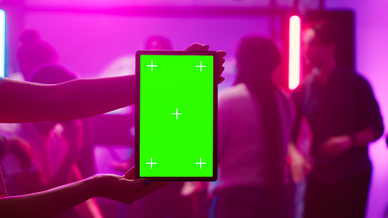 Woman holding greenscreen tablet at club and showing isolated display during dance party. Young funky adult using chroma key mockup template with blank copyspace on device screen.