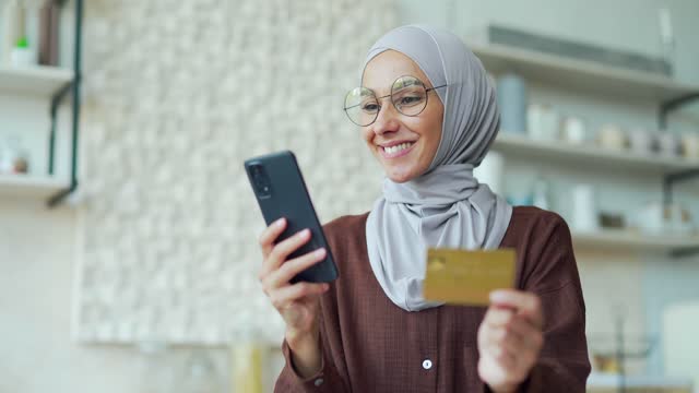 Young Muslim woman in hijab with glasses paying with credit card on smartphone in the kitchen Happy customer doing payments online shopping in internet store and receiving cashback indoors Easy pay