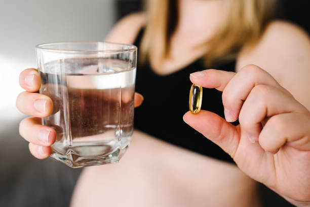 oméga 3. la femme enceinte prend des vitamines tous les jours et de l’eau en verre. les mains féminines tiennent des suppléments ou des vitamines. fille assise dans son lit à la maison. grossesse, traitement, médicaments concepts de soins de santé. - fatty acid photos et images de collection