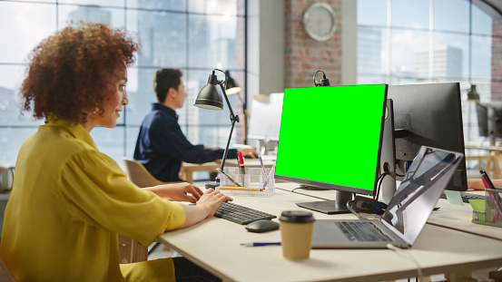 Employees Smiling While Working on Computers in Busy Modern Bright Office. Biracial Woman Designing Using Green Screen. Male Employee Working in the Background.