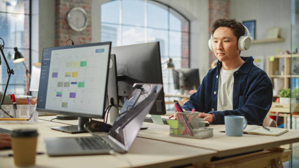 istock Portrait of an Asian Man Smiling While Using Computer and Headphones in Bright Modern Office. Communications Coordinator Planning a Project While Listening to Podcast. 1481164976