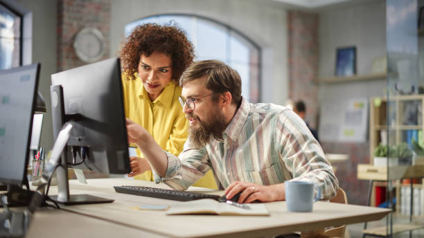 istock Portrait of Two Creative Colleagues Collaborating and Pointing at the Computer Screen in Modern Bright Office. Biracial Young Woman Discussing Work with Caucasian Male Assistant. 1481165438
