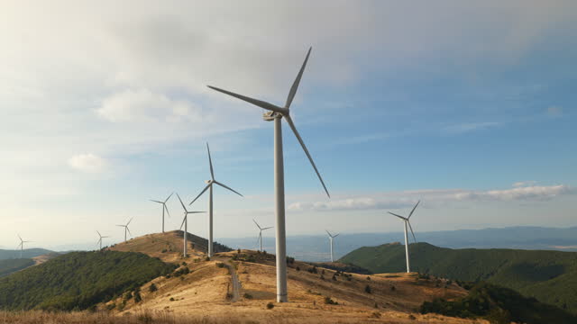 Large wind turbines with blades in mountains on peaks aerial view sunset blue sky wind park drone turn. Silhouettes of windmills, summer lenses. Alternative green energy