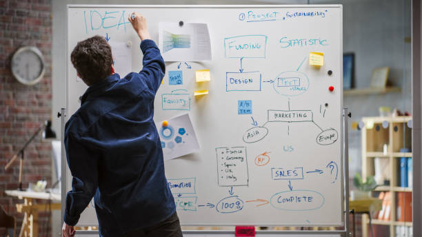 junger projektmanager, der diagramme und statistiken verwendet und auf ein whiteboard schreibt. spezialist für digitale inhalte beim brainstorming für eine marketingkampagne in einem büro. rückansichtsaufnahme. - co founder stock-fotos und bilder