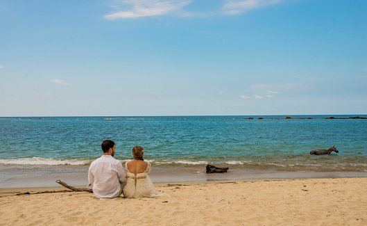 Young couple in white summer clothes sitting on the sand looking at the ocean in the afternoon.