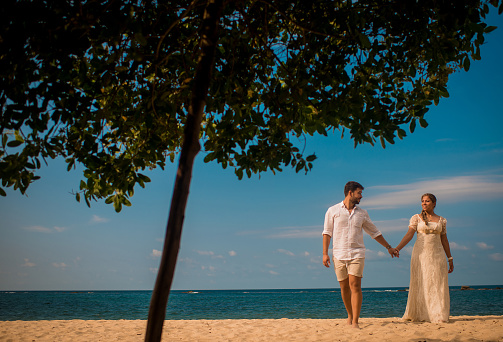 Couple holding hands in summer and walking on the beach in white costumes.