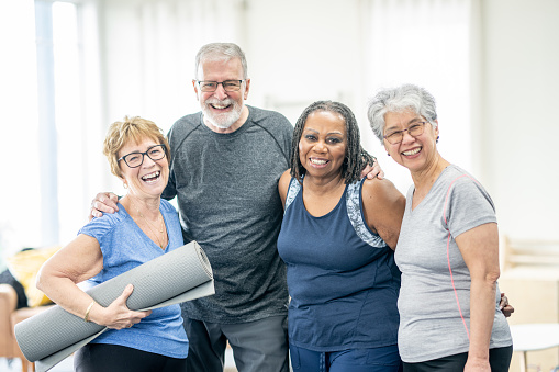 A small group of multi-ethnic seniors pose together for a portrait after a fitness class.  They are each dressed comfortably in athletic wear and are smiling.