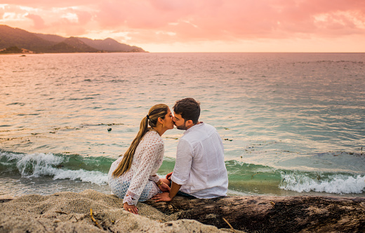 couple in love sitting on the shore of a beach while a red sunset is setting and kissing