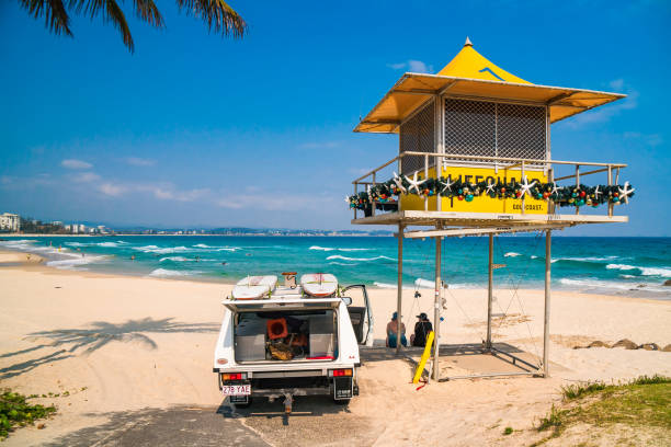 lifeguard truck with surfboards on the roof parked next to the yellow observation tower, and two women sitting in the shade of it. - gold coast australia lifeguard sea imagens e fotografias de stock