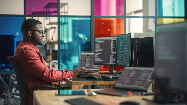 african american man writing lines of code on desktop computer with multiple monitors and laptop in creative office. male data scientist working on innovative online service for start-up company. - technicus stockfoto's en -beelden