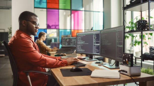 istock Black Man Writing Lines of Code On Desktop PC With Two Monitors and a Laptop Aside in Stylish Office. Professional Male Developer Programming Artificial Intelligence Software for Start-Up Company. 1481181825