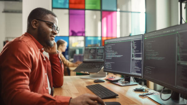 istock Black Man Writing Lines of Code On Desktop PC With Two Monitors and a Laptop in Stylish Office. Professional Male Developer Programming Artificial Intelligence Software for Start-Up Company. 1481181837