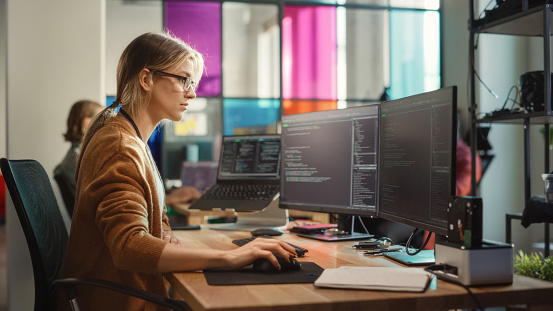 Caucasian Woman Coding on Desktop PC and Laptop Setup With Multiple Displays in Spacious Office. Female Junior Software Engineer Working on New Sprint of Mobile Application Development For Start-up.