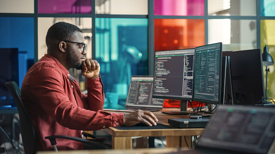 Male Senior Software Developer Writing Code On Desktop Computer With Two Displays Setup in Stylish Office. Black Man Working On Mobile Application For Online Service of Modern Start-up Company.