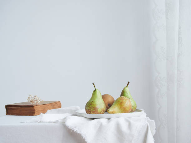 istock Simple minimalist indoor lifestyle composition: pears on a ceramic plate, next to an old book with dried flowers on top, on a table with white tablecloth next to the net curtain. Light wall with copy space in the background. 1481193084