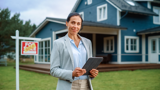 Portrait of an Intelligent Real Estate Agent in Smart Suit Holding Tablet Computer, Looking at Camera and Smiling. Multiethnic real estate agent Standing in Front of Residential Property.