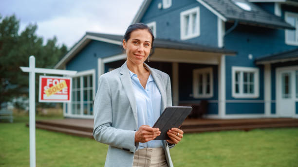 Portrait of an Intelligent Real Estate Agent in Smart Suit Holding Tablet Computer, Looking at Camera and Smiling. Multiethnic real estate agent Standing in Front of Residential Property. stock photo